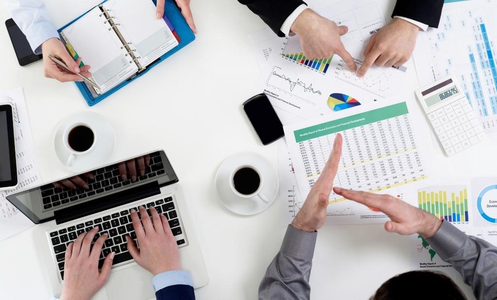 Visão aérea de uma mesa de escritório na qual um grupo de pessoas está trabalhando em equipe. Há papéis, gráficos e um laptop. Duas pessoas apontam para um relatório financeiro, enquanto outra digita no computador.