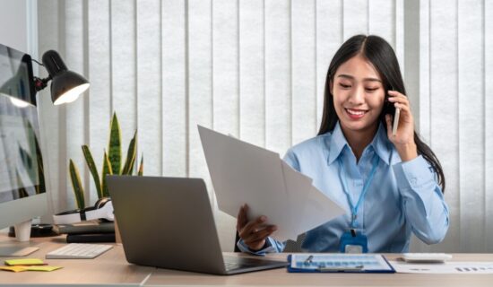 Mulher sorridente vestindo uma camisa azul e usando um crachá, sentada à mesa de trabalho. Ela está ao telefone e segurando um documento. Na mesa, há um laptop, uma luminária, alguns papéis e uma planta decorativa ao fundo