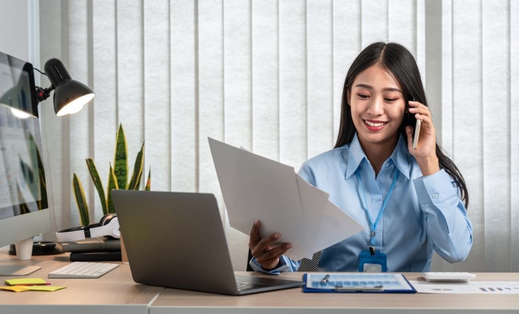 Mulher sorridente vestindo uma camisa azul e usando um crachá, sentada à mesa de trabalho. Ela está ao telefone e segurando um documento. Na mesa, há um laptop, uma luminária, alguns papéis e uma planta decorativa ao fundo
