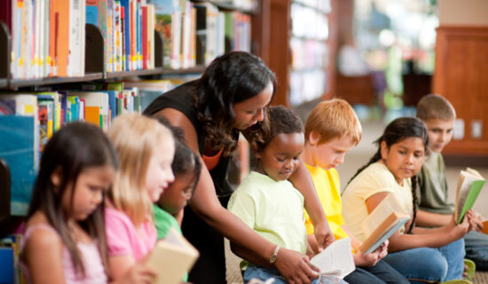 Roda de alunos com a professora indicando um trecho de um livro para um aluno.