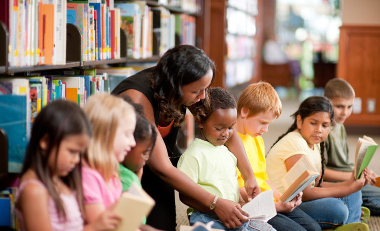 Roda de alunos com a professora indicando um trecho de um livro para um aluno.