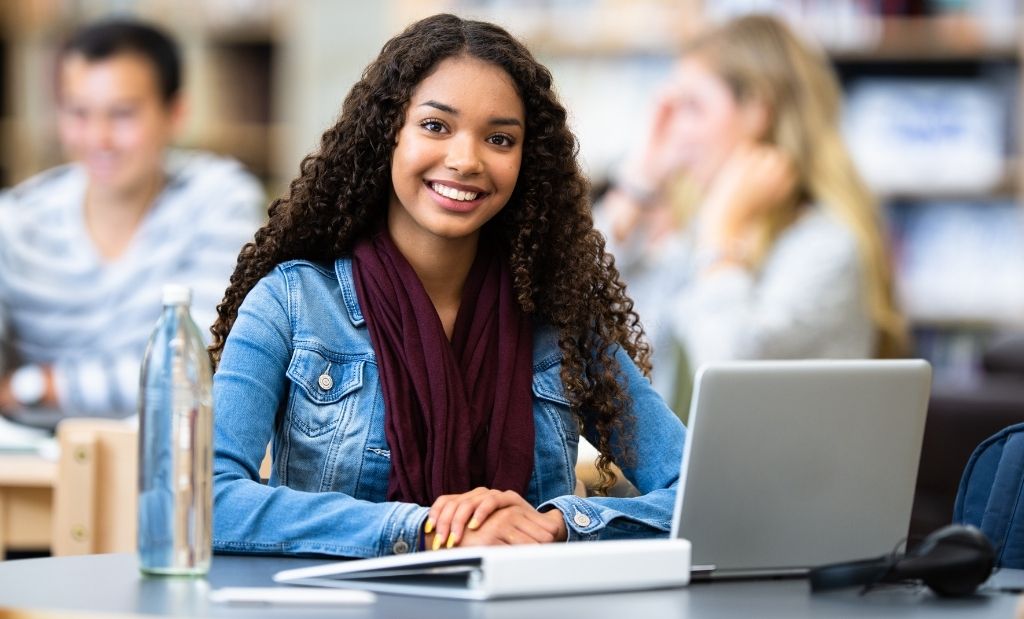 jovem estudante sorrindo enquanto usa um laptop em um ambiente escolar moderno.