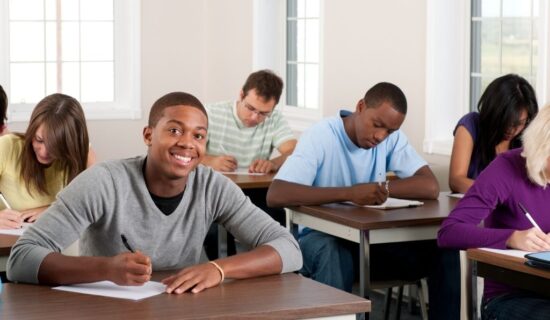 grupo de alunos concentrados escrevendo em uma sala de aula, um deles sorrindo confiante.