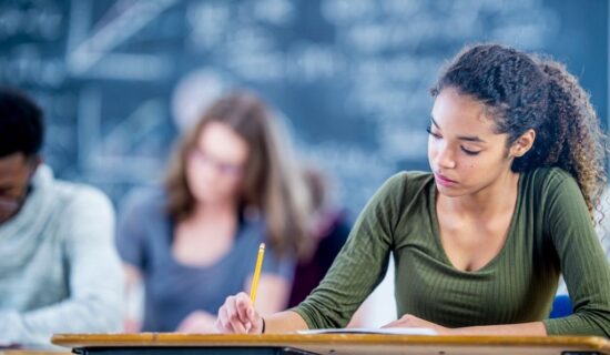Uma jovem estudante está sentada em uma mesa de aula, concentrada, enquanto escreve com um lápis em uma folha. Ao fundo, outros alunos estudam em uma sala com lousa.