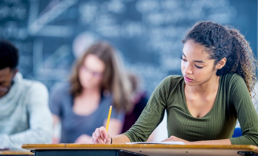 Uma jovem estudante está sentada em uma mesa de aula, concentrada, enquanto escreve com um lápis em uma folha. Ao fundo, outros alunos estudam em uma sala com lousa.